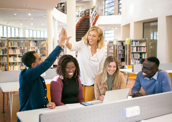 Cheerful diverse group of students collaborating in a library, celebrating with a high five.